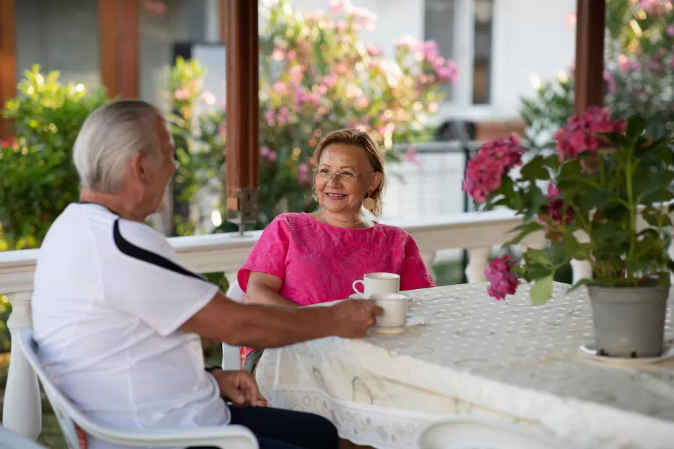 Elderly woman in pink shirt sitting at table on porch across from elderly man in white shirt. A pot of pink flowers sits on the table.